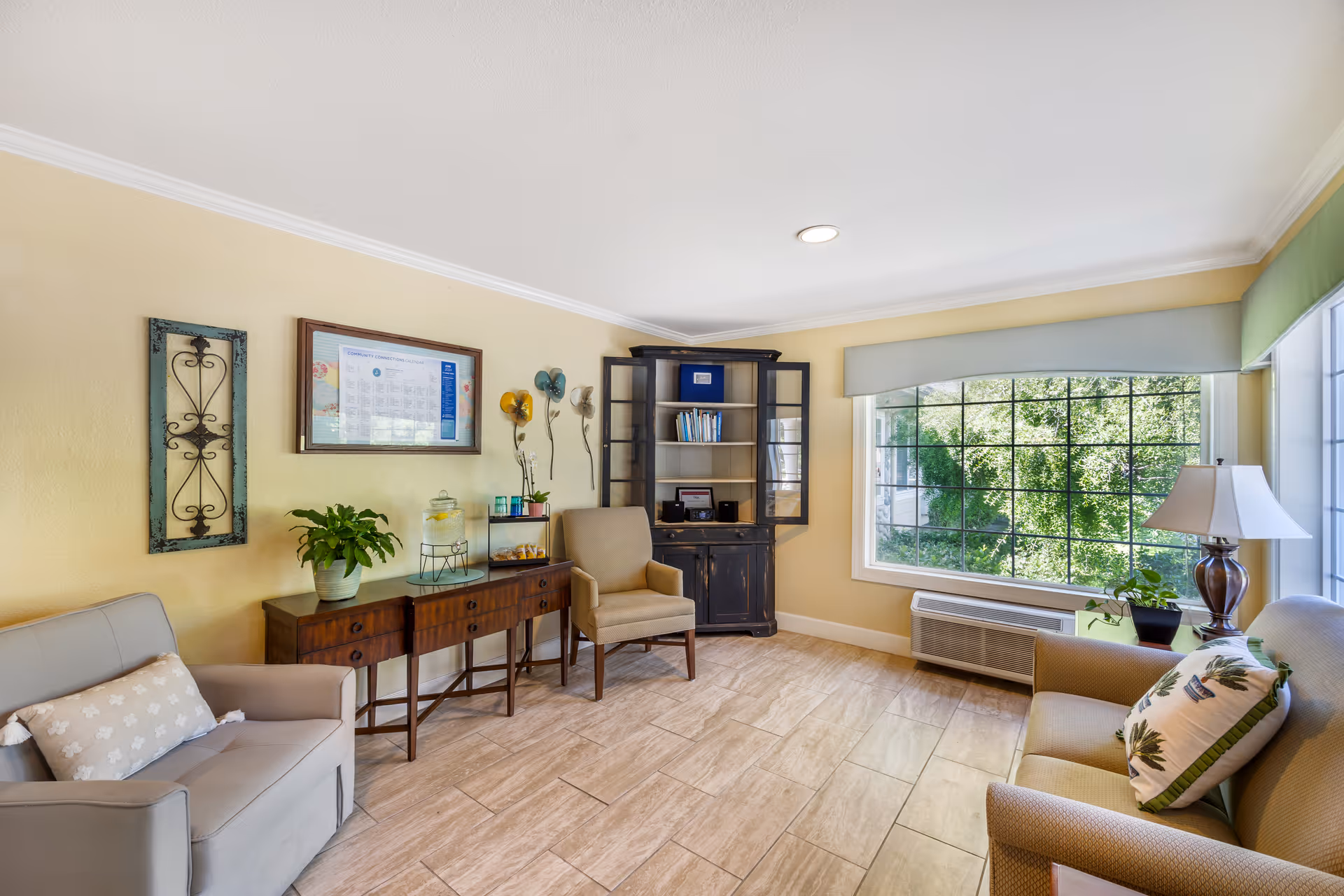 A bright and cozy living room area with beige walls and tiled floor. The room features a large window with a view of green trees outside, a beige armchair, a gray loveseat with a decorative pillow, a wooden sideboard with plants and a water dispenser, a black cabinet with glass doors containing books, and a table lamp on a side table.