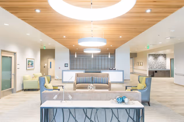 Bright and modern senior living facility common area with comfortable seating including a sofa and armchairs arranged around a coffee table. The ceiling features wooden slats and large circular light fixtures. The space has light-colored walls and flooring, with artwork on the walls and a reception desk in the background.