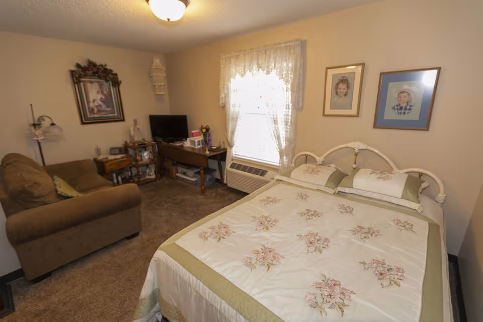 A cozy furnished bedroom with a bed, sofa, desk and TV beside a window with lace curtains.