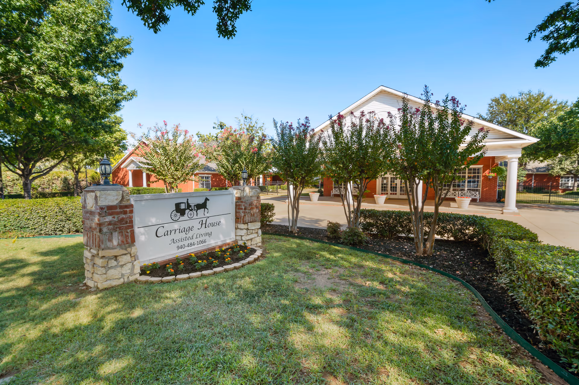 Exterior view of Carriage House Assisted Living of Denton facility with a sign in the foreground surrounded by flowers and greenery, trees lining the driveway, and a brick building with white columns in the background under a clear blue sky.