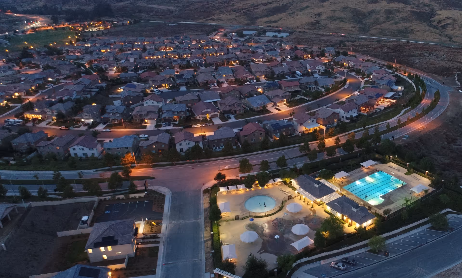Aerial dusk view of a residential neighborhood with an illuminated community pool and surrounding houses.