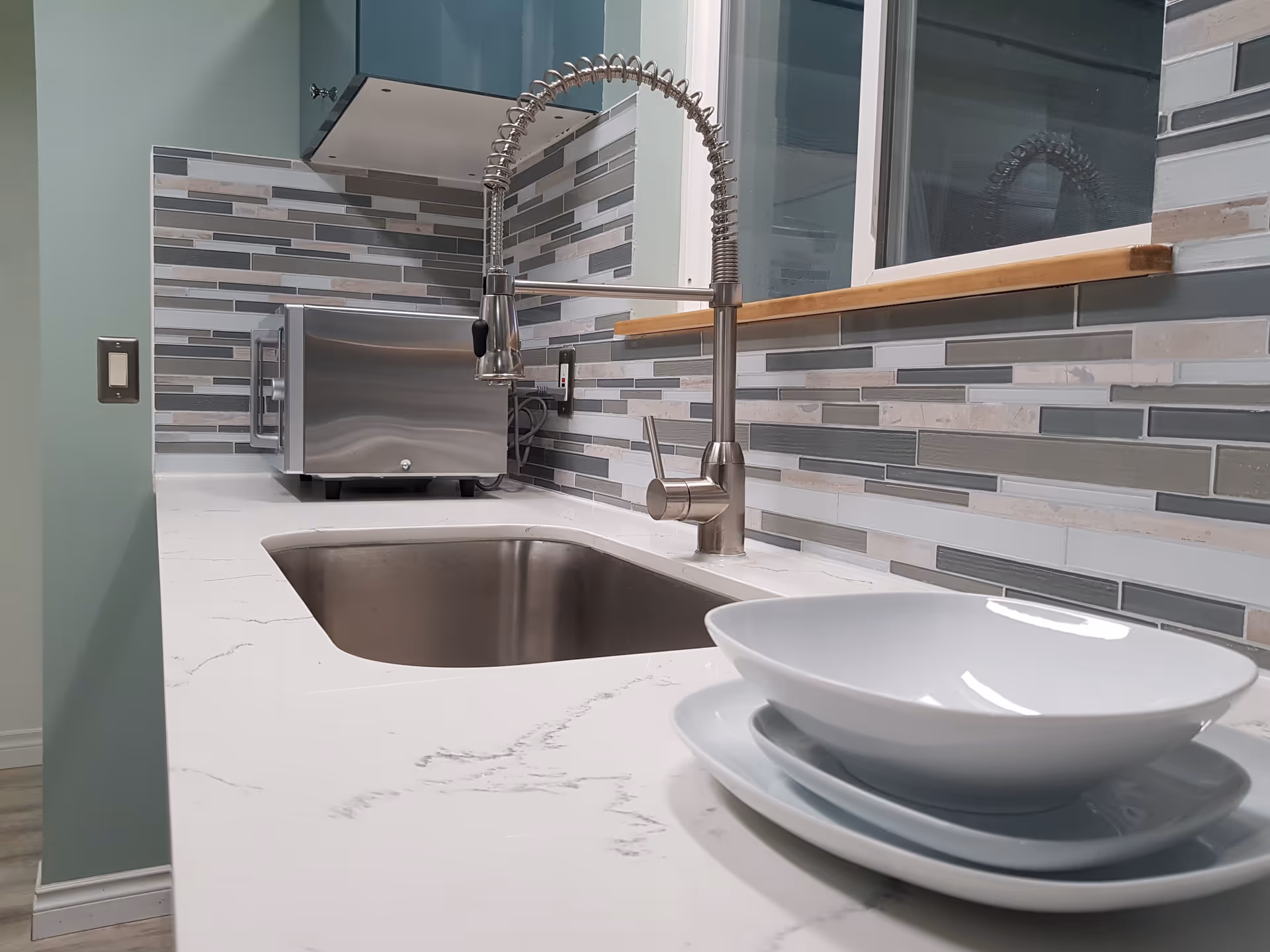 Modern kitchen countertop with a stainless steel sink and a flexible spring faucet. There is a stainless steel microwave on the counter against a tiled backsplash with various shades of gray and beige. In the foreground, there are stacked white plates and a bowl.