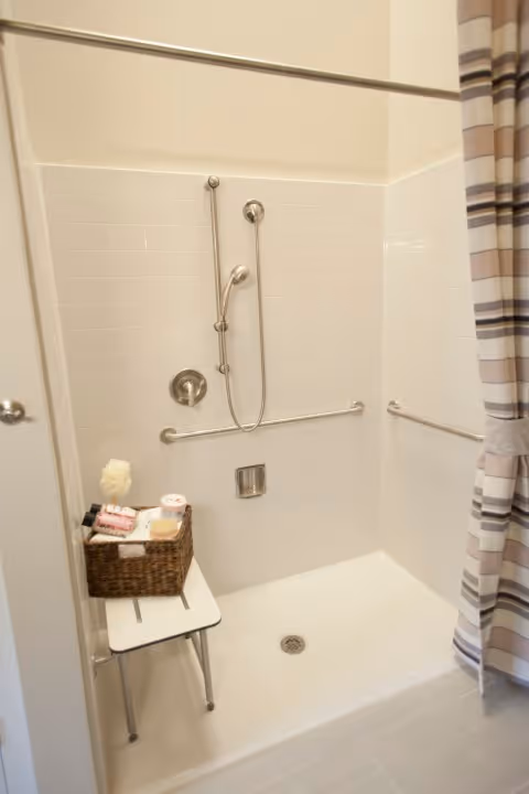 A walk-in shower with white tiled walls and a handheld showerhead mounted on a vertical bar. There are two horizontal grab bars on the walls for support. A small white shower bench holds a basket containing toiletries and a loofah. A striped shower curtain is partially visible on the right side.