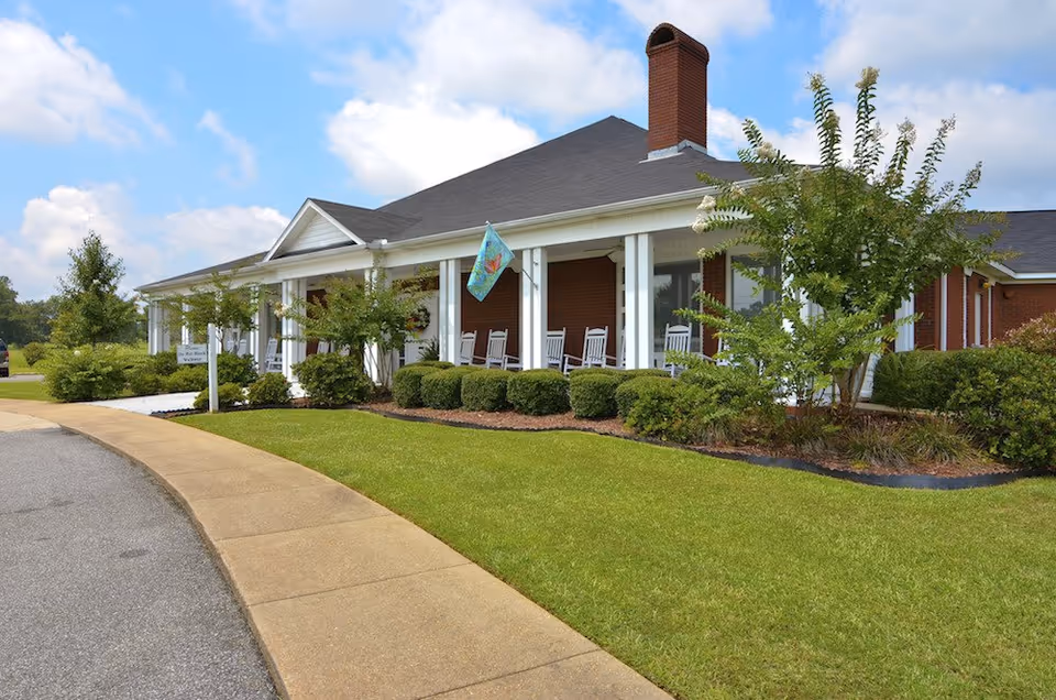 Brick one-story senior living facility with a wrap porch, rocking chairs, and manicured lawn.
