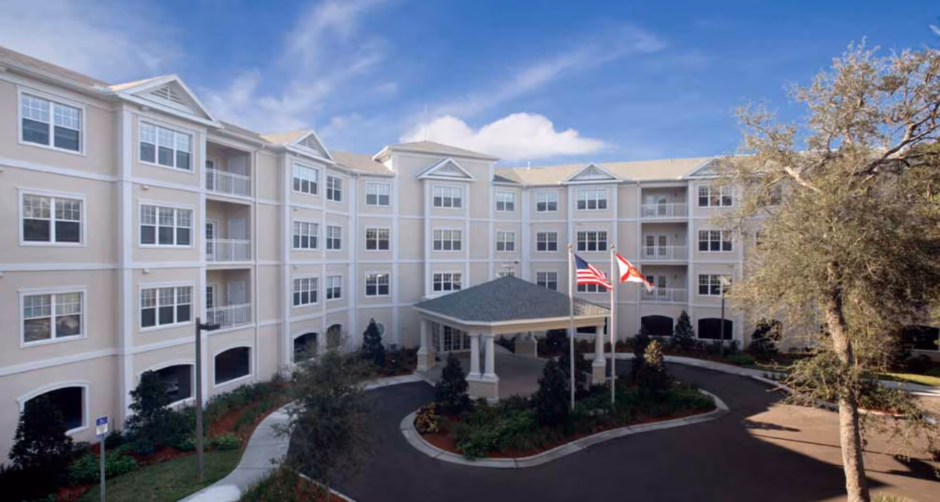 Exterior view of a multi-story senior living facility building with beige walls and white trim. The building has multiple windows and balconies. In front of the building is a circular driveway with a covered entrance supported by columns. Two flagpoles display the American flag and a state flag. There are landscaped bushes and trees around the driveway under a partly cloudy blue sky.