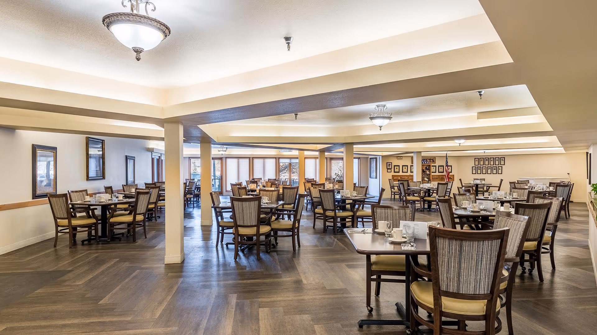 A spacious dining room with multiple round and square tables, each surrounded by wooden chairs with cushioned seats. The tables are set with cups, glasses, and napkins. The room has a light-colored ceiling with recessed lighting and large windows along the back wall allowing natural light to enter.