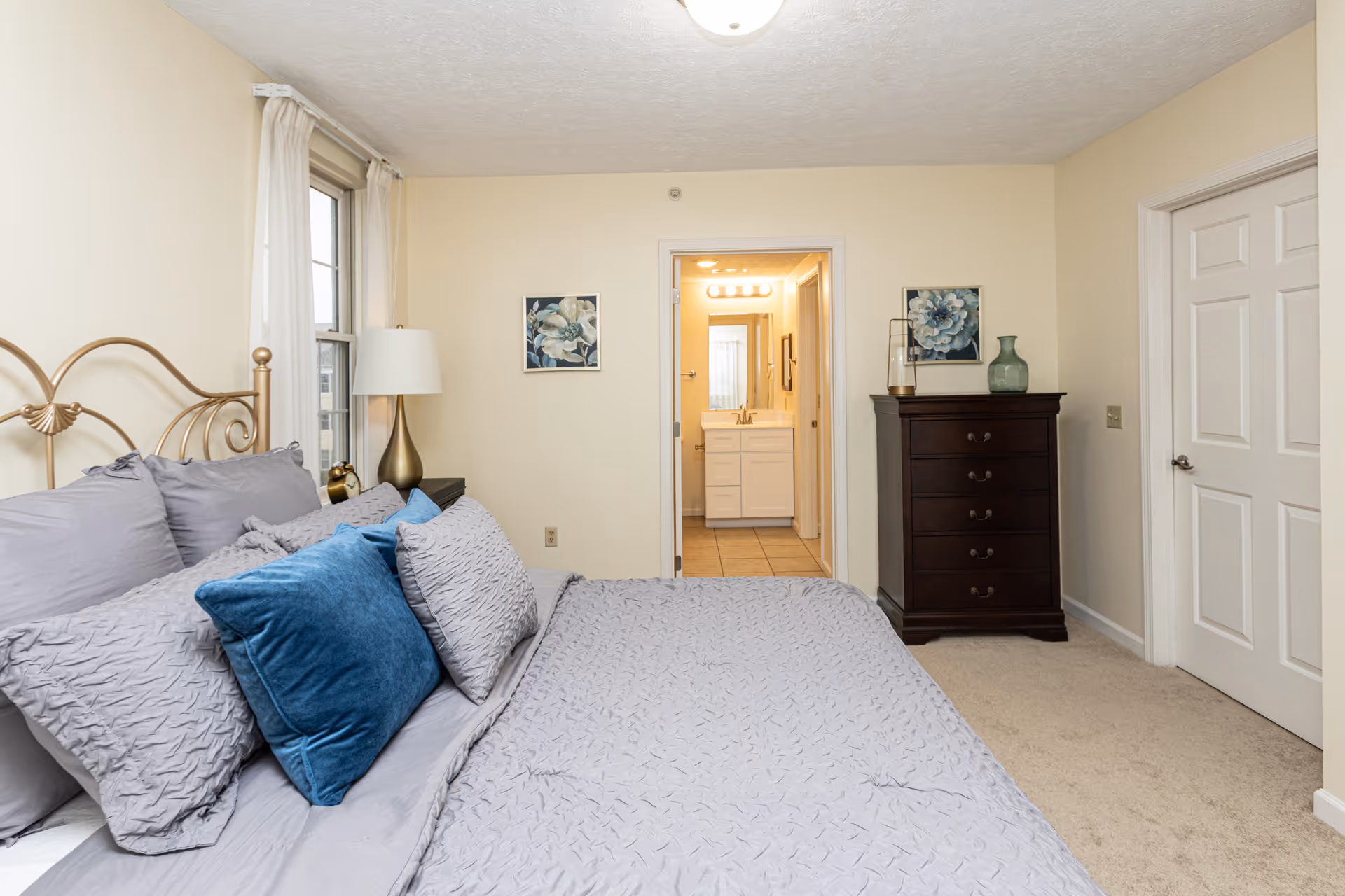 A bedroom with a neatly made bed featuring gray bedding and blue accent pillows. To the left of the bed is a window with white curtains and a gold lamp on a nightstand. On the wall opposite the bed are two framed floral pictures, a dark wooden dresser with decorative items on top, and a door leading to a bathroom with a white vanity and mirror. Another closed door is visible on the right side of the room.