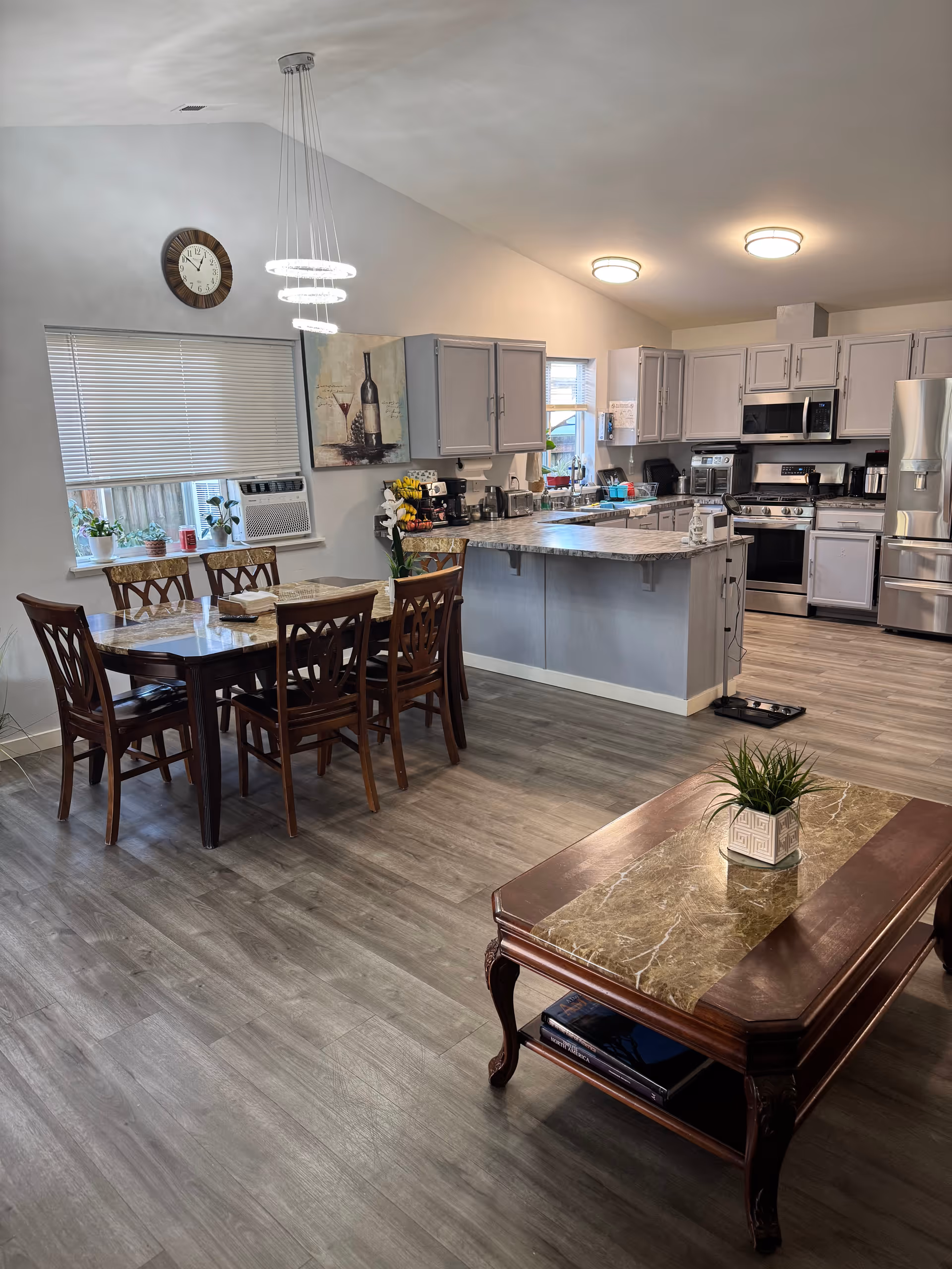 A spacious and modern kitchen and dining area with light gray cabinets, stainless steel appliances, and a marble countertop island. There is a wooden dining table with six chairs near a window with blinds and an air conditioning unit. A decorative wall clock and a painting of a wine bottle and glass hang on the wall. In the foreground, there is a wooden coffee table with a marble top and a small potted plant.