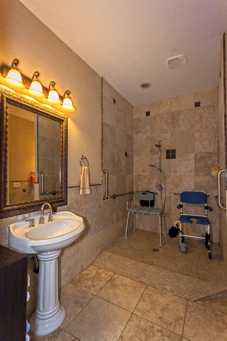 A bathroom with beige tiled walls and floor, featuring a white pedestal sink with a framed mirror above it and a towel ring with a white towel. The shower area has two shower chairs, one green and one blue, with grab bars installed on the walls for support.