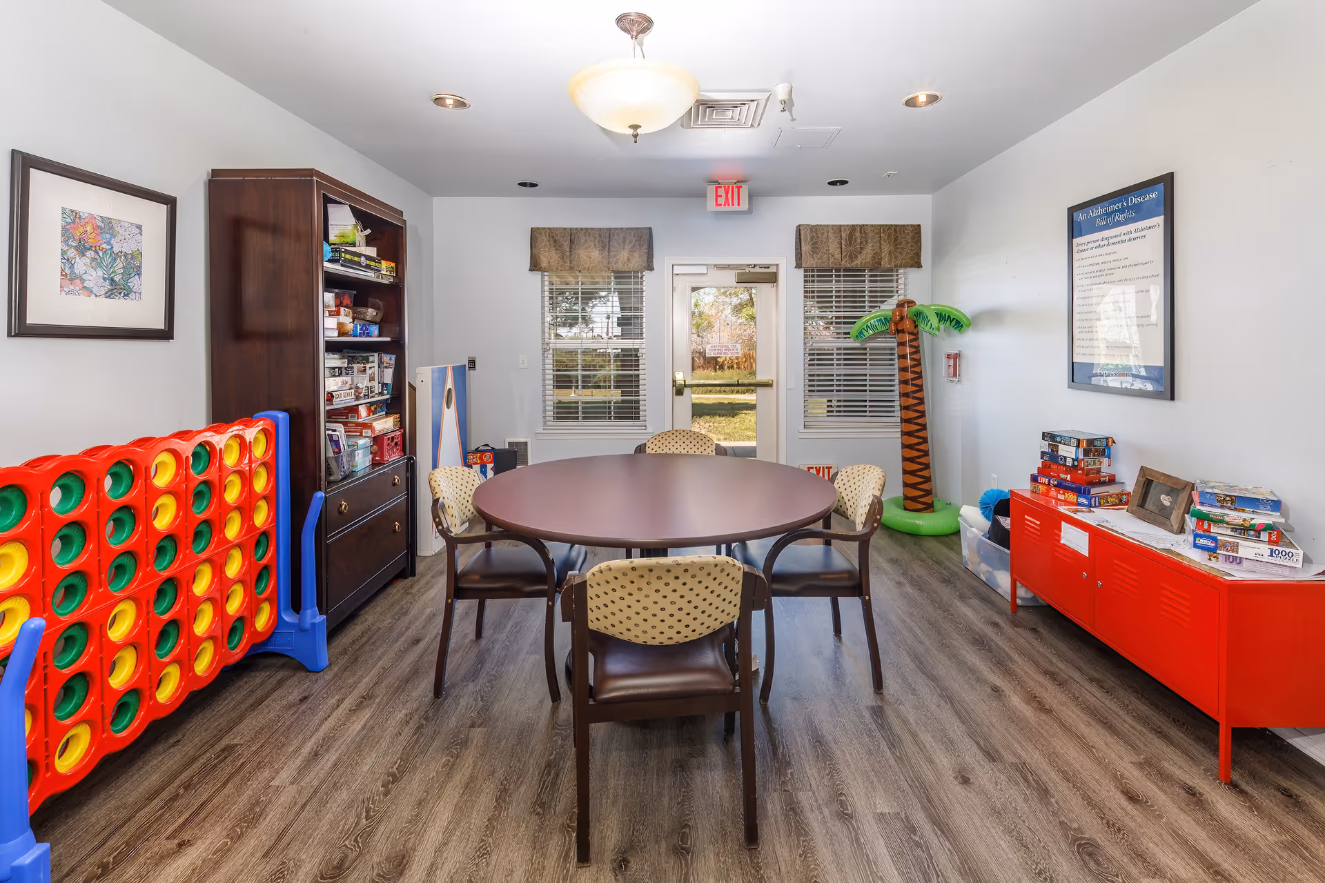 A bright activity room with a round table surrounded by four chairs. On the left side, there is a large Connect Four game and a tall dark wooden bookshelf filled with board games and puzzles. On the right side, a red cabinet holds more board games and puzzles, with an inflatable palm tree decoration nearby. Two windows with valances and a glass door leading outside are visible at the back of the room. The floor is wood laminate, and the walls are white with framed artwork and an informational poster.