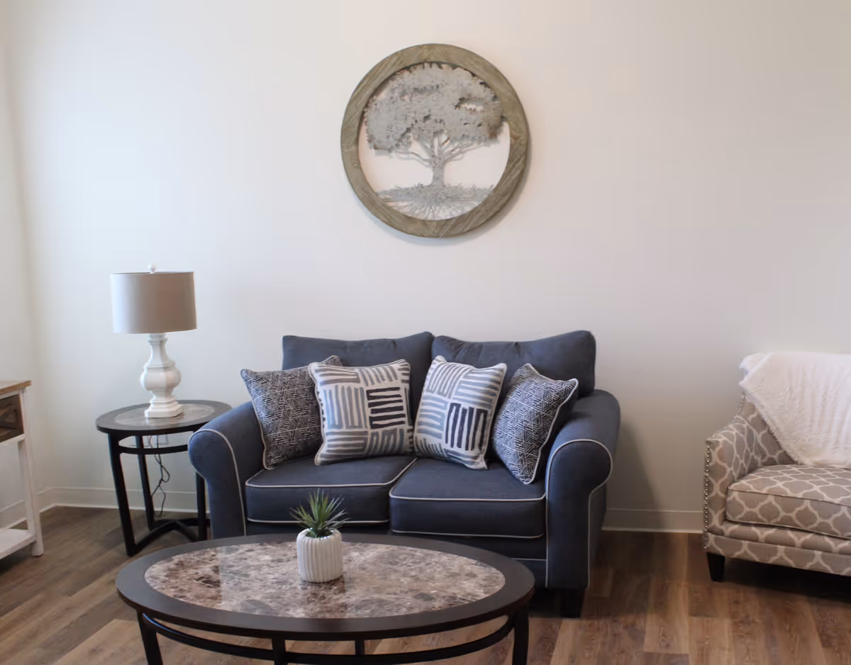 Small living room with a blue loveseat topped with patterned pillows, a marble-top coffee table, side table with a lamp, and round tree wall art.