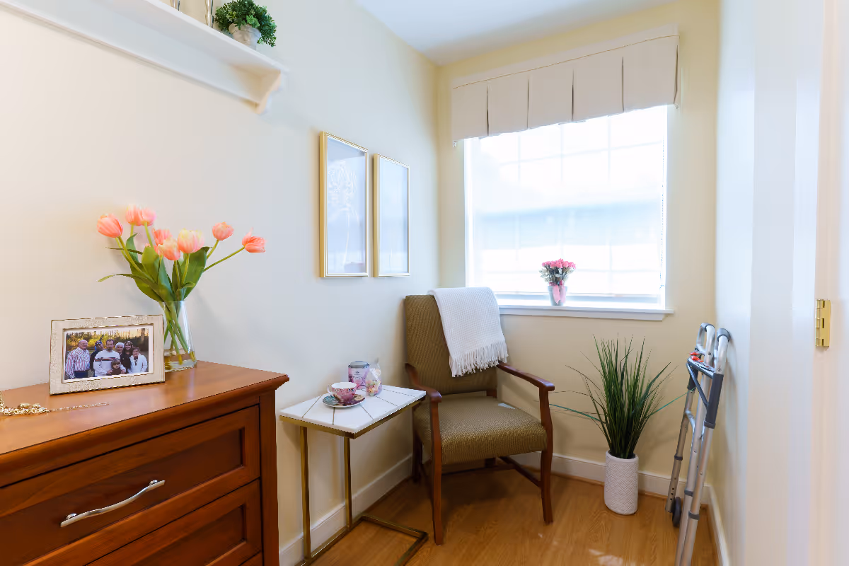 A cozy corner of a senior living facility room with a wooden dresser topped with a framed family photo and a vase of pink tulips. Next to it is a small side table with a teacup and saucer, and a comfortable armchair with a white throw draped over the back. A window with a beige valance lets in natural light, and a potted plant and a walker are placed near the wall.