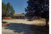 Single-story brick residential building with a gravel parking area, trees, and a few parked cars.