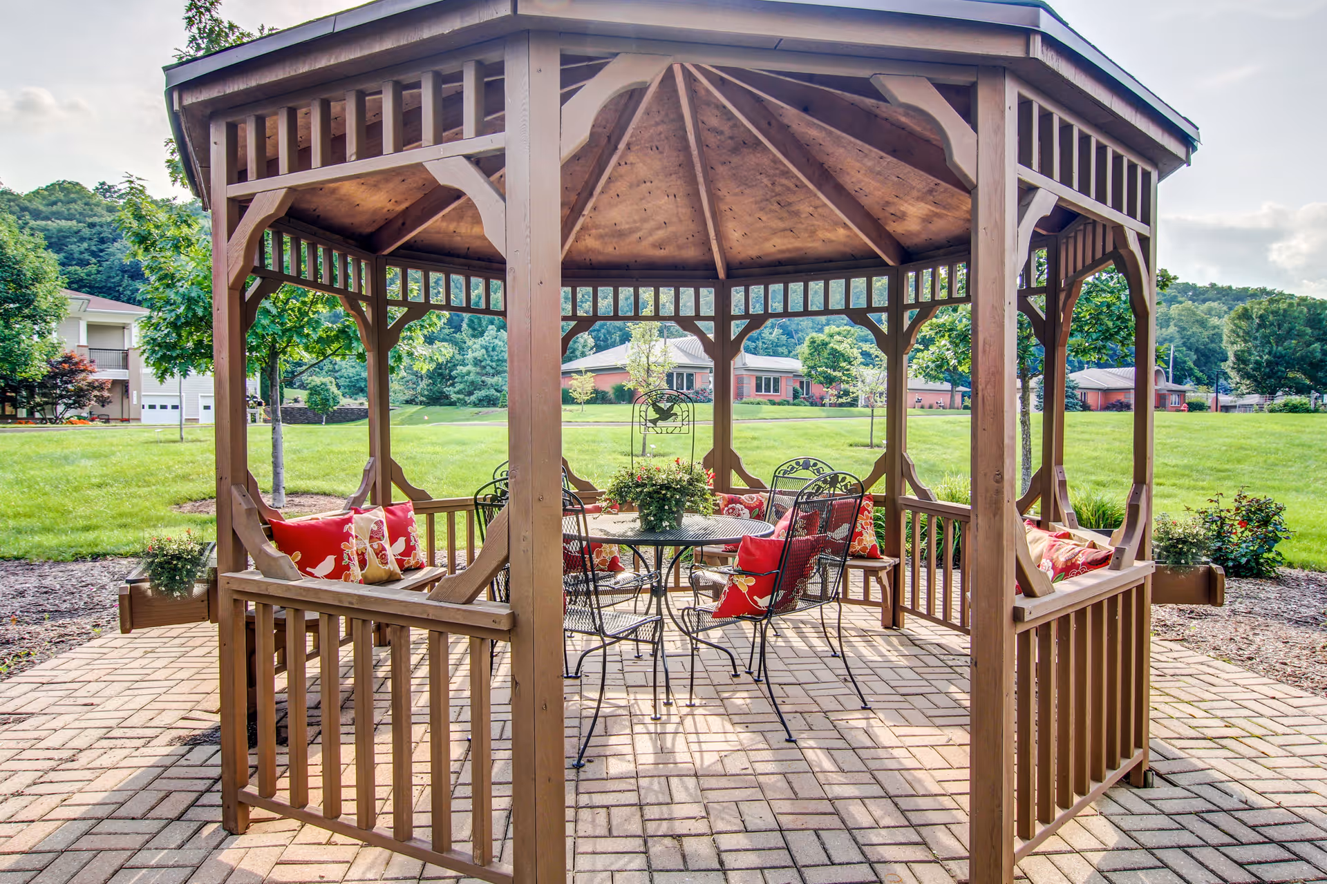 A wooden gazebo with a pitched roof situated on a paved patio area. Inside the gazebo, there is a round metal table surrounded by four metal chairs with red cushions and pillows. The gazebo is set in a green outdoor space with trees, grass, and buildings in the background under a partly cloudy sky.