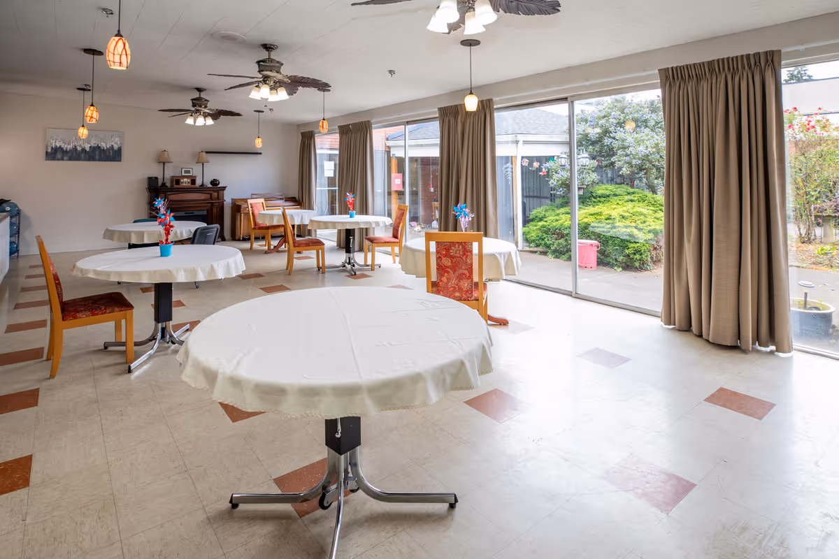 A bright dining room with round tables covered in white tablecloths and wooden chairs with red patterned cushions. Large sliding glass doors on one side provide a view of a garden with green bushes and flowers. The room has ceiling fans with lights, pendant lights, and beige curtains framing the glass doors. There is a piano and some lamps against the far wall.
