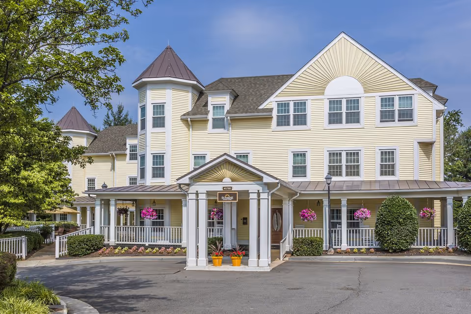 Front exterior of a yellow three-story senior living building with a covered entrance, turret, porch, and hanging flower baskets.