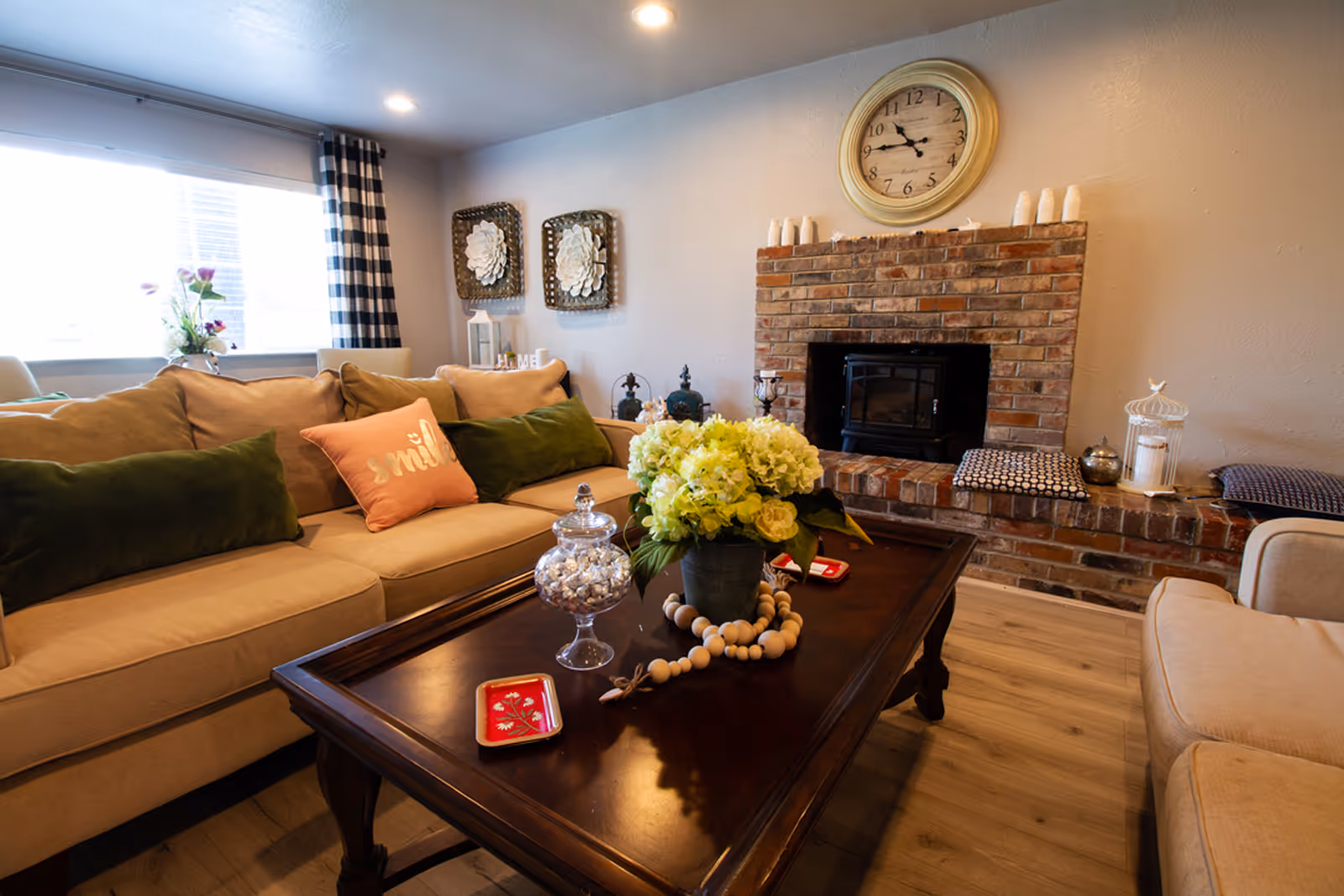 Cozy living room with beige sofas adorned with green and pink pillows, a dark wooden coffee table with a flower arrangement and decorative items, a brick fireplace with a clock above it, and a window with checkered curtains letting in natural light.