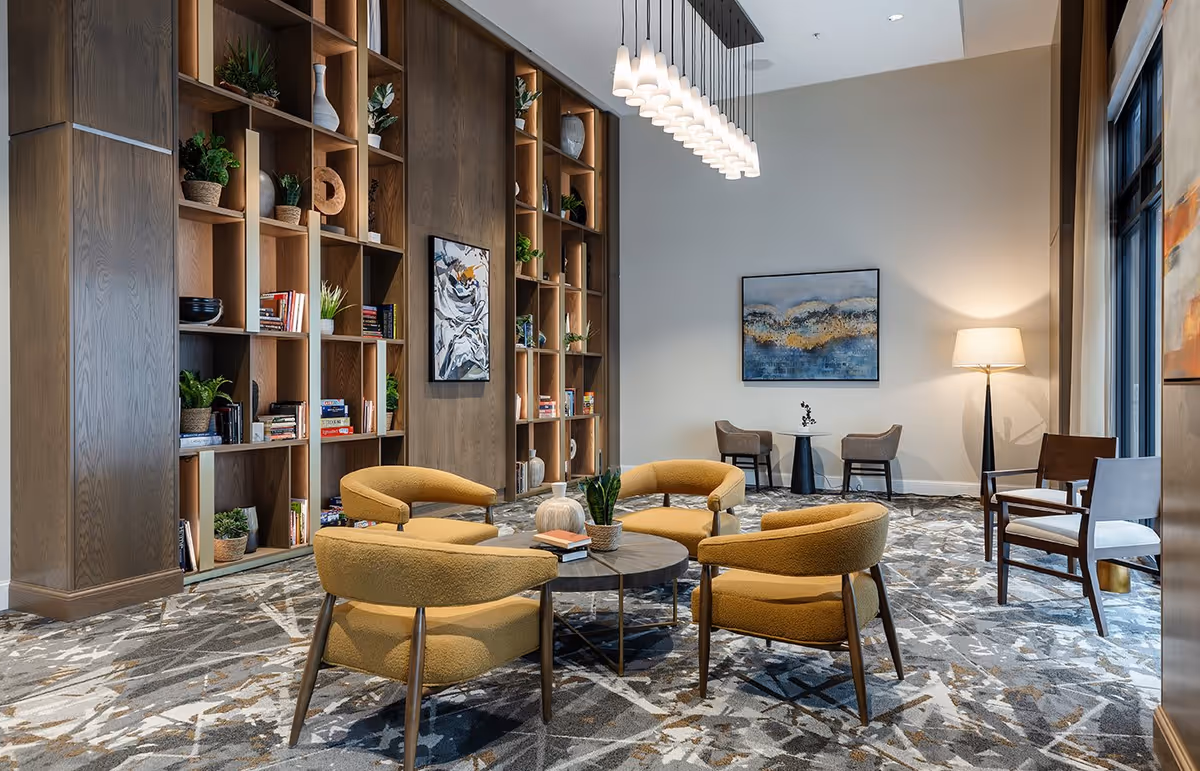 A modern lounge area with four mustard-yellow upholstered chairs arranged around a round wooden coffee table with books and plants. Behind the seating area is a tall wooden bookshelf filled with books, plants, and decorative items. The room features a patterned carpet, a floor lamp, two additional chairs with a small round table, and abstract artwork on the walls.