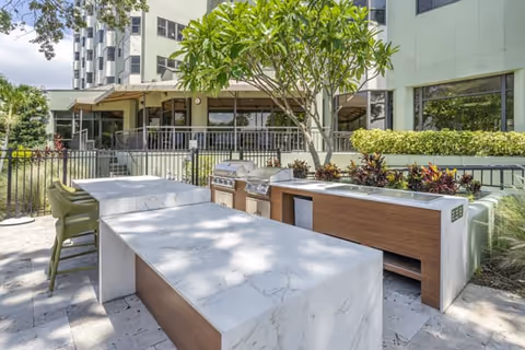 Outdoor patio area at Brookdale Bayshore featuring modern marble countertops with built-in grills and seating, surrounded by greenery and adjacent to the building with large windows.