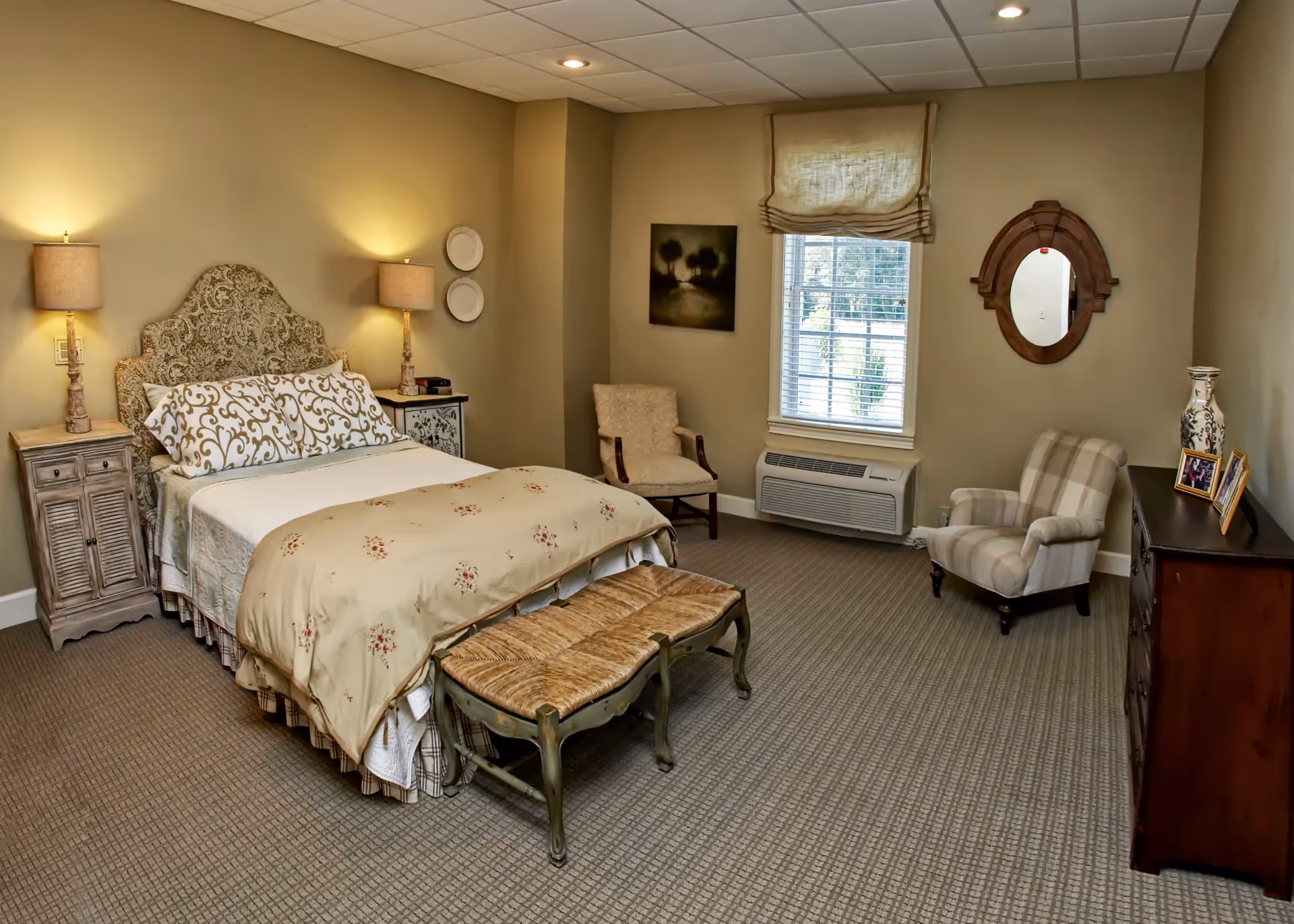 A cozy bedroom with a bed featuring a patterned headboard, two bedside tables with lamps, a bench at the foot of the bed, two armchairs, a window with blinds and a Roman shade, a wooden dresser with framed photos and a decorative vase, a round mirror on the wall, and a painting beside the window.