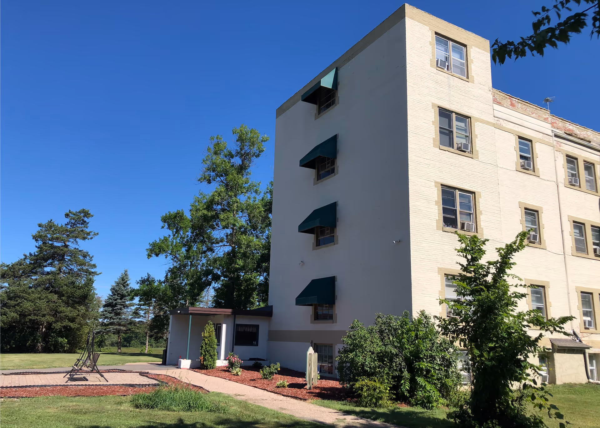 Exterior view of a multi-story beige brick building with green window awnings, surrounded by green trees and bushes under a clear blue sky. There is a paved walkway and a small patio area with a swing in the grassy yard.