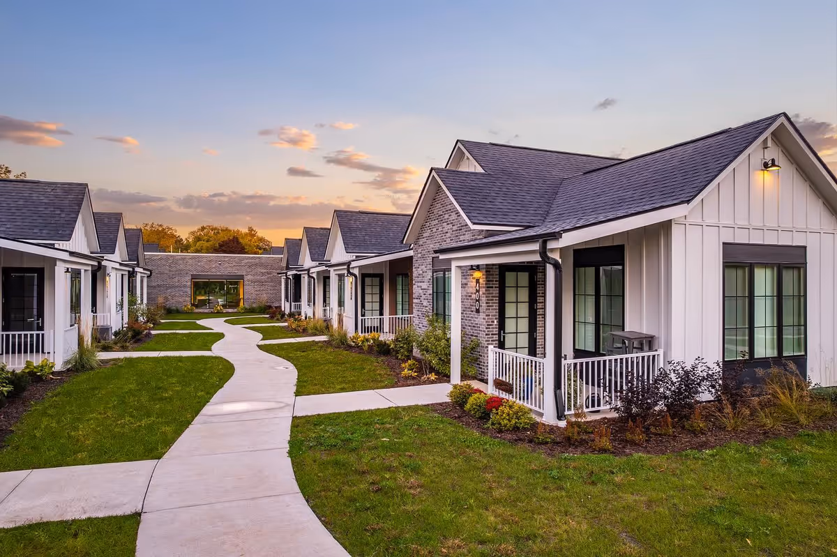 A pathway winds through a green lawn flanked by modern single-story residential buildings with white siding, black-framed windows, and small porches. The sky is partly cloudy with a warm sunset glow.