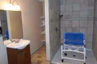Bathroom with sink, mirror, and blue shower chair in a tiled shower.