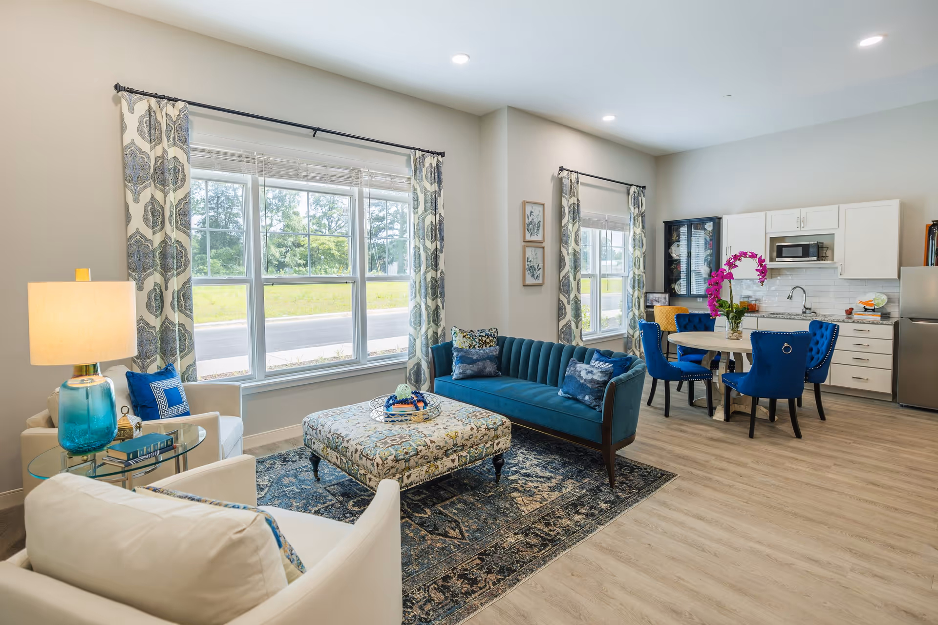 Bright open-plan living area with a teal sofa, patterned ottoman, blue dining chairs, and a small kitchen under large windows.
