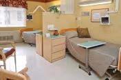 Shared patient bedroom with two hospital-style beds, bedside storage, overbed tables, chairs, wall-mounted monitors, and a window.