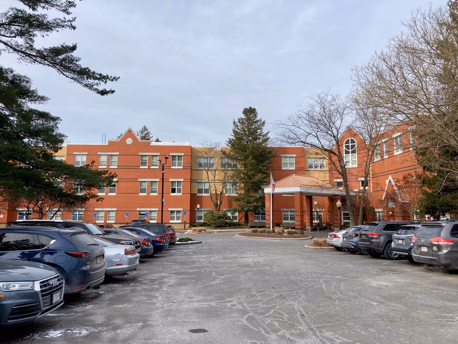 Exterior view of Spaulding Nursing & Therapy Center, a three-story brick building with a covered entrance and several windows. The parking lot in front is filled with parked cars, and there are trees on both sides of the building under a cloudy sky.