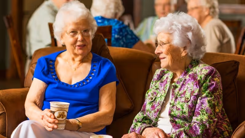 Two elderly women sitting on a brown couch in a common area, engaged in conversation. One woman is wearing a blue top and holding a cup, while the other is wearing a floral jacket. Other elderly individuals are visible in the background, seated and interacting.
