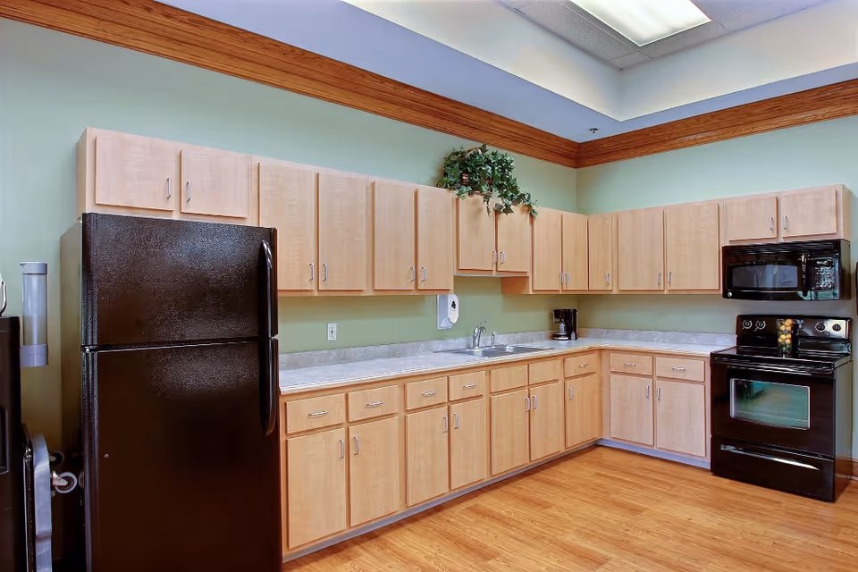 A clean and modern kitchen with light wood cabinets, a black refrigerator, black stove with oven, black microwave, and a coffee maker on the countertop. The walls are painted light green with a wooden trim near the ceiling, and the floor is a light wood laminate.