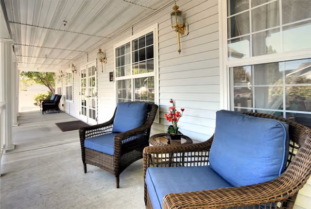 A covered outdoor porch area with wicker chairs featuring blue cushions and small round tables with decorative flowers. The porch has white siding walls, multiple windows, and brass wall-mounted lanterns. Trees and greenery are visible in the background.