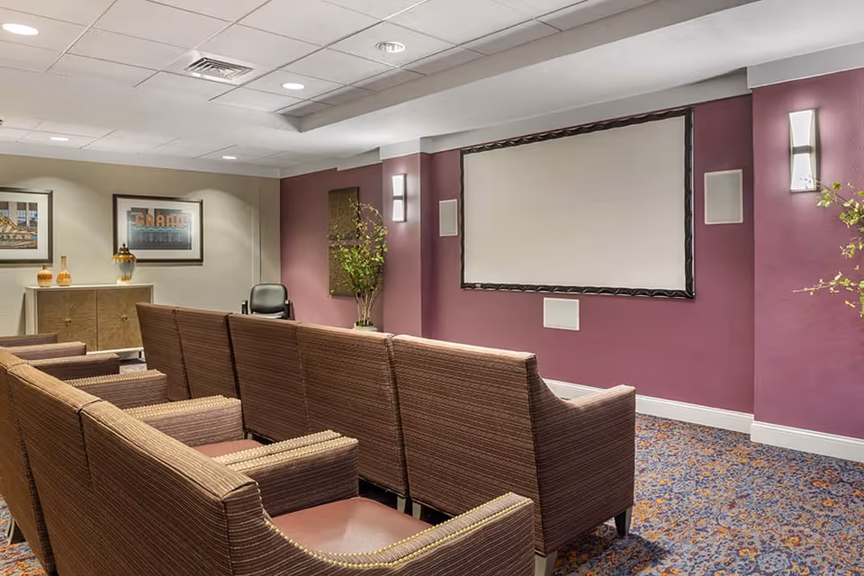 A small theater room with rows of brown upholstered chairs facing a large blank projection screen mounted on a purple wall. The room has patterned carpet, wall sconces, framed artwork, and a cabinet with decorative items.