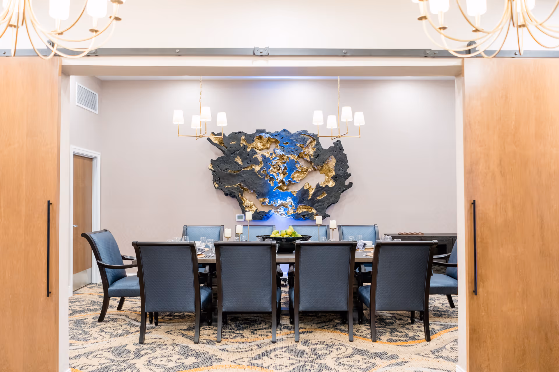A formal dining room with a long rectangular table surrounded by eight blue upholstered chairs. Above the table hang two modern chandeliers with white lampshades. On the wall behind the table is a large abstract art piece in black, gold, and blue colors. The room has a patterned carpet and light-colored walls, with wooden sliding doors partially open to reveal the space.