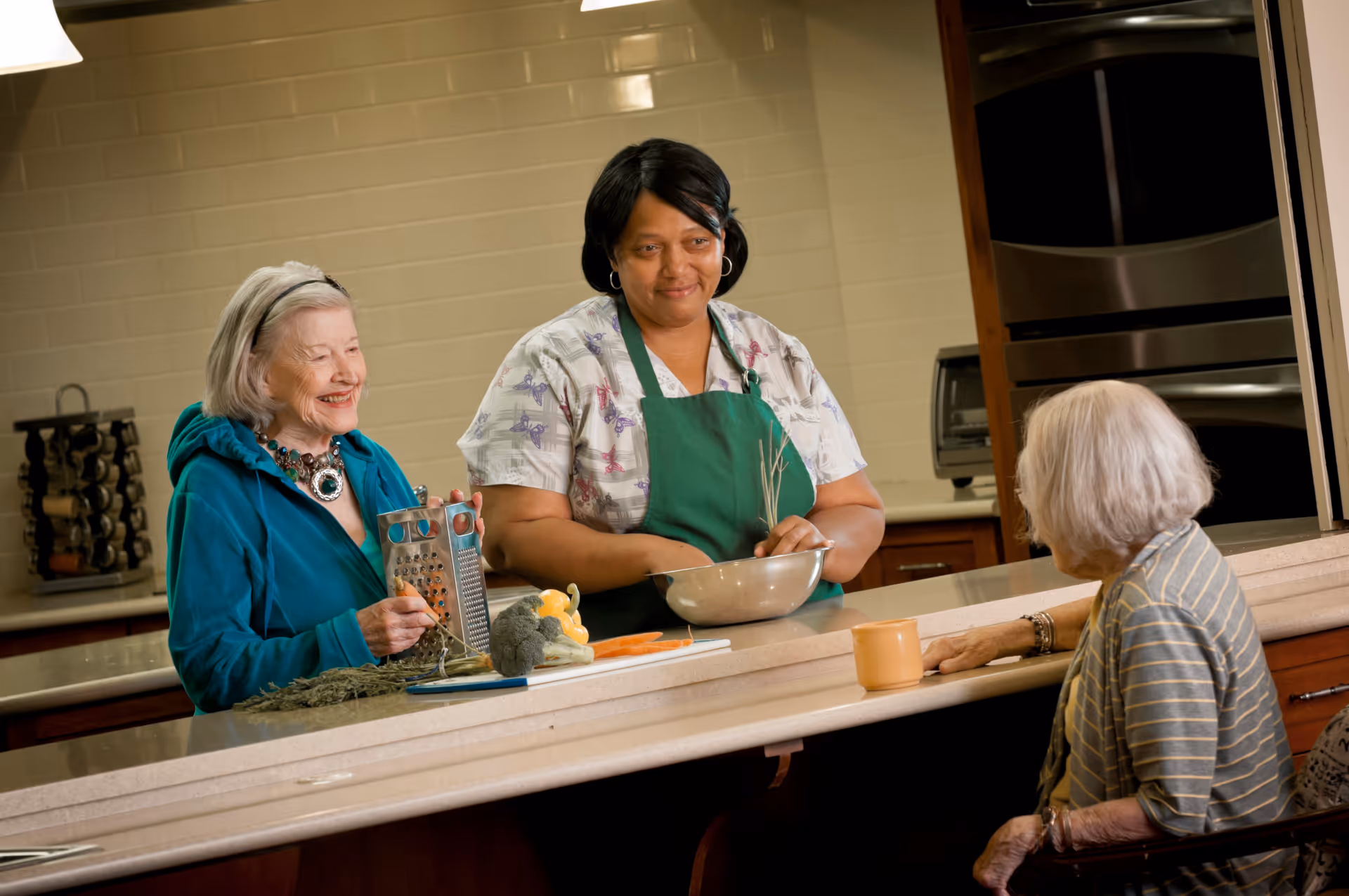 Three women in a kitchen setting; one woman in a green apron is mixing something in a bowl, an elderly woman in a teal jacket is holding a grater and smiling, and another elderly woman with white hair is sitting at the counter with a mug in front of her, engaging in conversation.