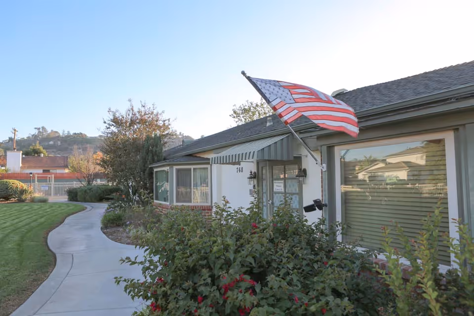 Front entrance of a single-story senior living building with an American flag, curved walkway, and landscaped shrubs.
