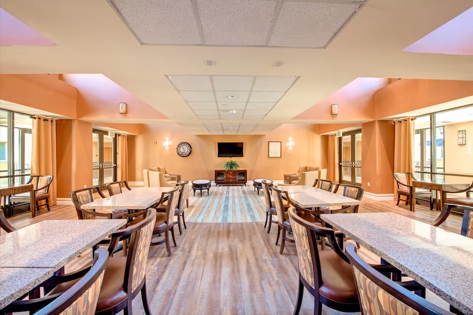 Bright communal dining area with multiple tables and chairs leading to a seating lounge and wall-mounted TV.