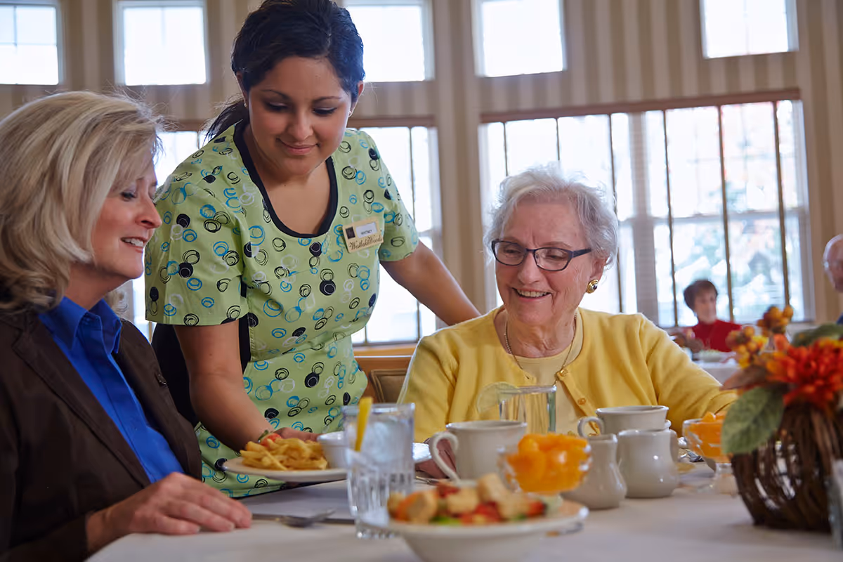 A care staff member serves meals to two older women seated at a bright dining room table with cups and bowls.