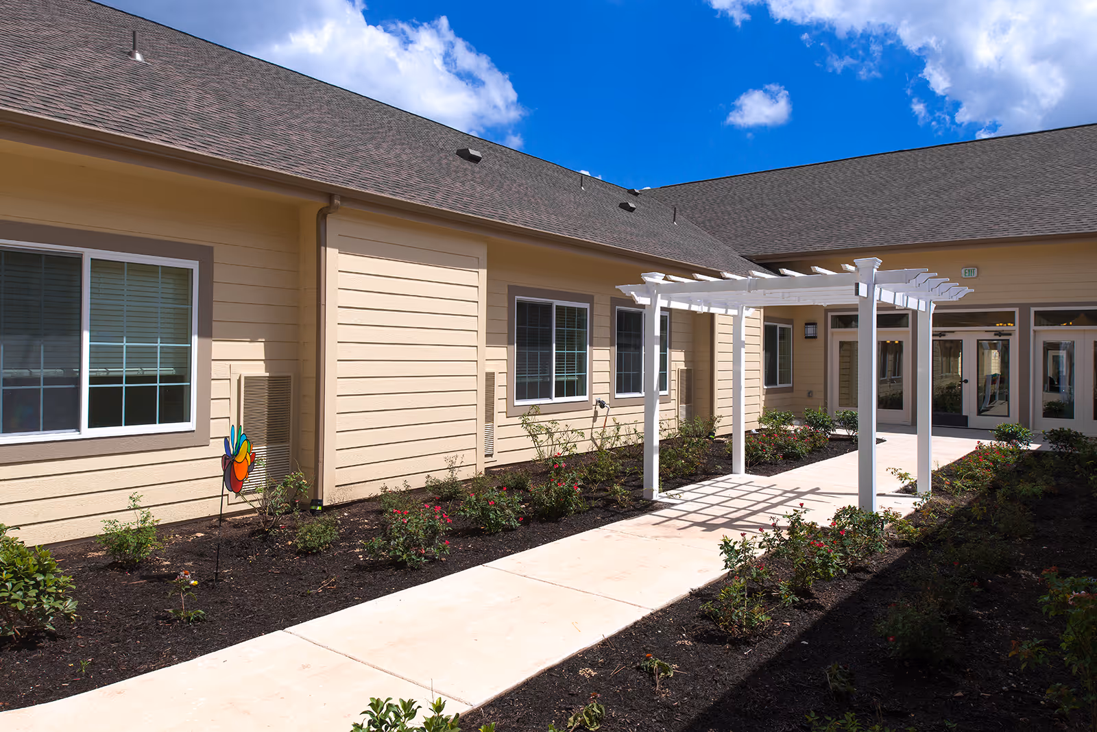Outdoor courtyard area at The Monarch at Cedar Park featuring a beige building with multiple windows, a white pergola over a concrete walkway, and landscaped flower beds with small bushes and plants under a partly cloudy blue sky.