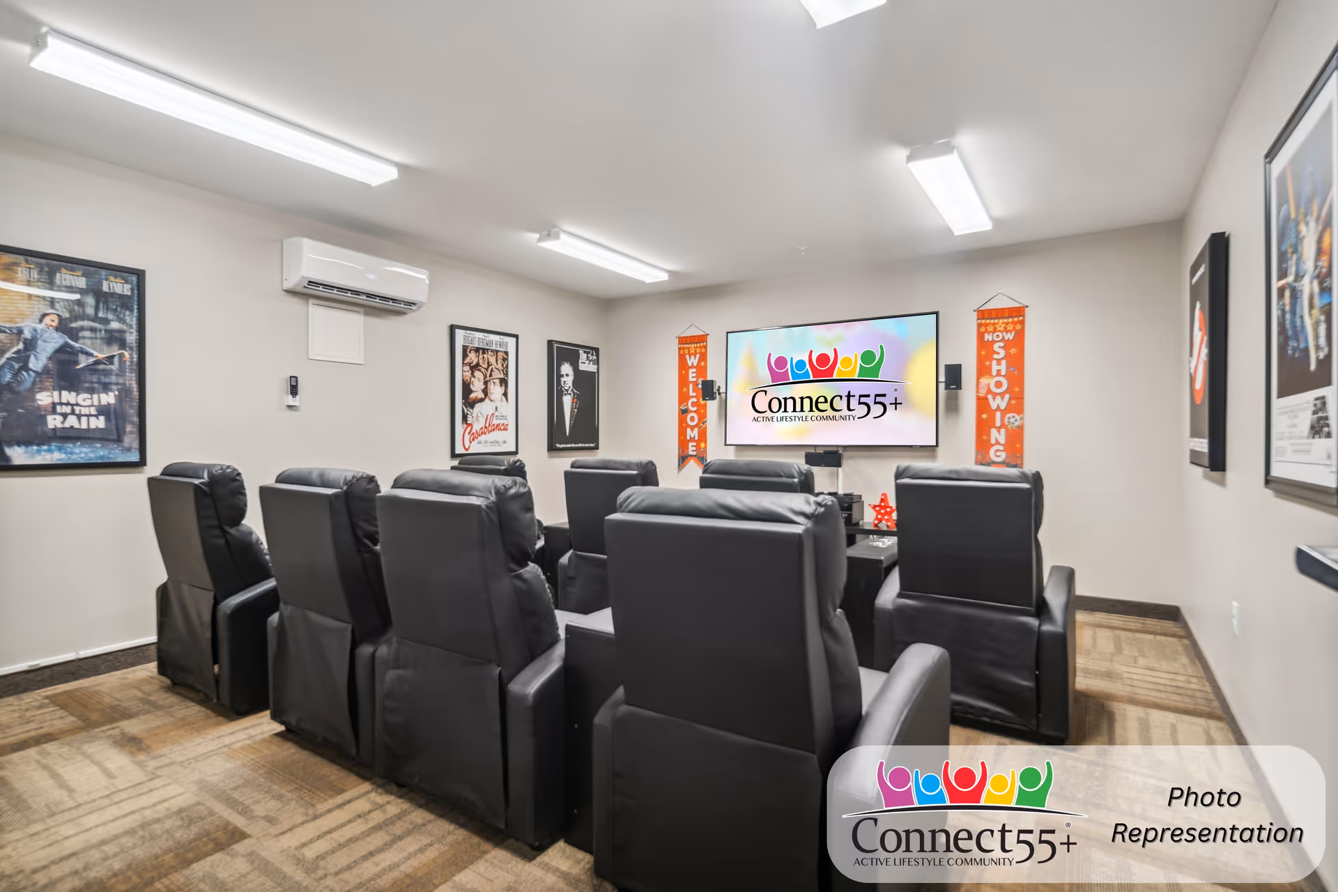 A small movie theater room with two rows of black leather recliner chairs facing a large flat-screen TV mounted on the wall. The TV screen displays the Connect55+ logo. The walls are decorated with classic movie posters including Singin' in the Rain, Casablanca, The Godfather, and Ghostbusters. There are also two vertical banners on either side of the TV that say 'WELCOME' and 'NOW SHOWING'. The room has bright overhead fluorescent lighting and a carpeted floor.