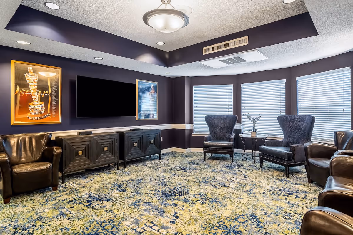 A cozy living room with dark purple walls and a patterned carpet in shades of blue and green. The room features a flat-screen TV mounted on the wall above two black cabinets, flanked by two framed pictures. There are several leather armchairs arranged around the room, with three large windows covered by white blinds letting in natural light. A small side table with a vase and flowers is placed between two armchairs near the windows.
