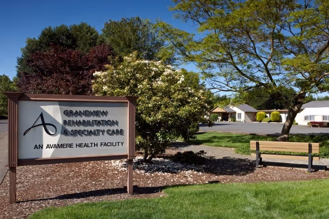 Outdoor view of the Grandview Rehabilitation & Specialty Care facility sign with trees, a bench, and buildings in the background under a clear blue sky.