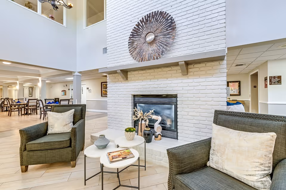A cozy senior living common area with two green armchairs featuring beige pillows facing a white brick fireplace. On the fireplace mantel is a decorative round mirror with a sunburst design. In front of the chairs are three small white nesting tables with decorative items including a book, a vase, and small plants. In the background, there is a dining area with tables and chairs, and the space has a bright, open layout with light-colored flooring and walls.