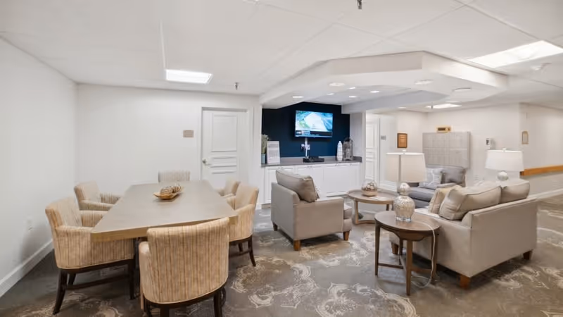A cozy common area in a senior living facility featuring a rectangular wooden table with six upholstered chairs on the left, and a seating area on the right with two beige armchairs, a beige sofa, two round wooden side tables with decorative lamps, and a round coffee table. A flat-screen TV is mounted on a dark accent wall above white cabinetry. The room has patterned carpet flooring and white walls with a drop ceiling.