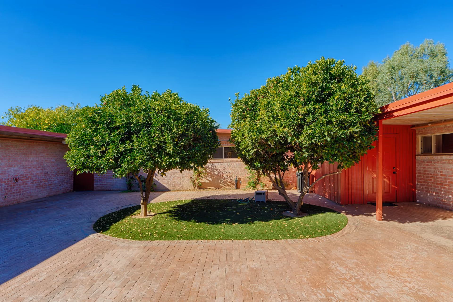 A sunny outdoor courtyard area with two leafy green trees planted in a small patch of grass surrounded by brick paving. The courtyard is enclosed by brick walls and features a red door and red wooden beams supporting a roofed area on the right side. The sky is clear and blue.