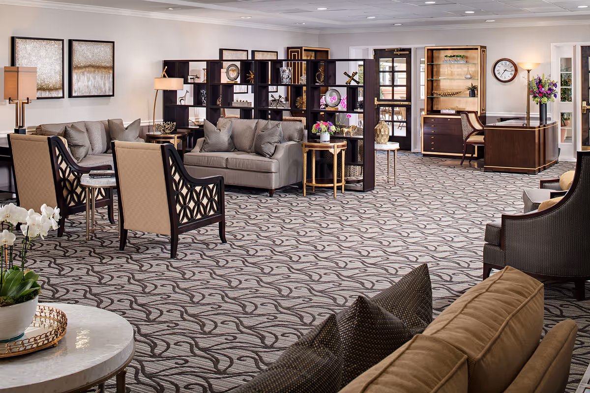A spacious and elegantly furnished senior living common area with patterned carpet, multiple seating arrangements including sofas and armchairs, decorative shelving with ornaments, a desk with a chair, wall clock, and floral arrangements. The room is well-lit with lamps and natural light from glass doors.