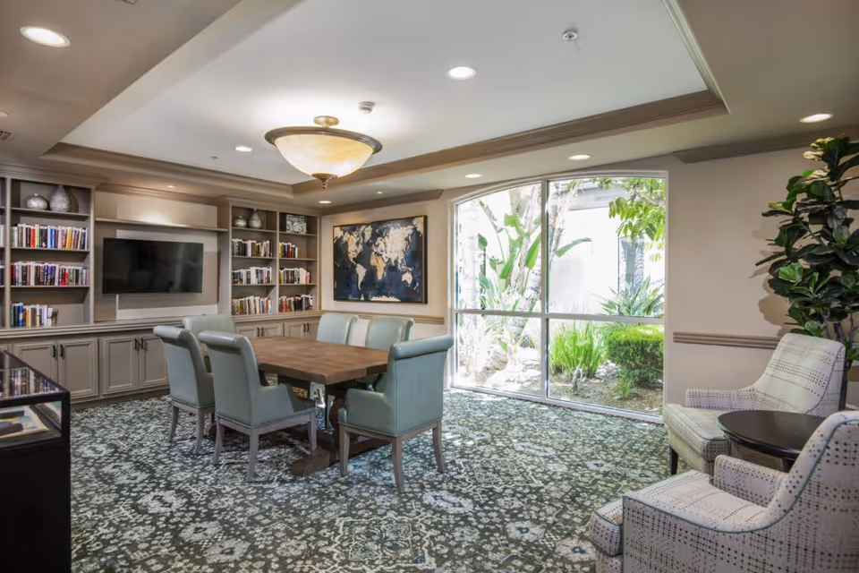 A well-lit common room with a large wooden table surrounded by six upholstered chairs in the center. Built-in bookshelves and cabinets line the back wall, with a flat-screen TV mounted in the middle. A large world map artwork hangs on the wall next to a large window that looks out onto greenery. Two patterned armchairs and a small round table are positioned near the window. The room has a patterned carpet and a ceiling light fixture.