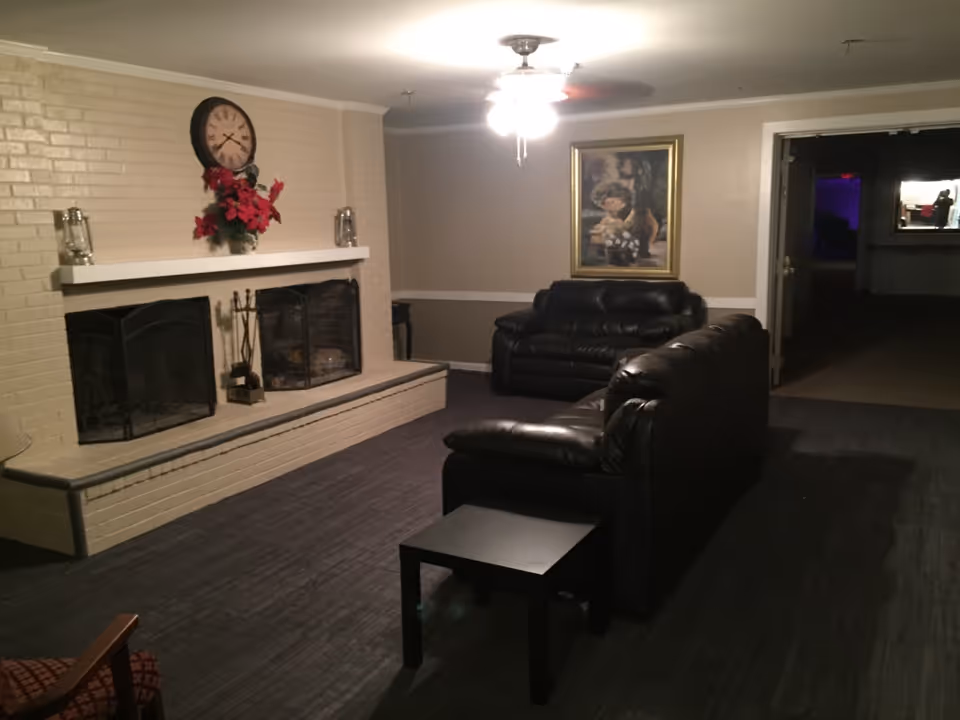 A cozy living room area with two black leather sofas facing a beige brick fireplace. The fireplace has a white mantle decorated with a clock, red flowers, and lanterns. A ceiling fan with lights is on, and a framed painting hangs on the wall behind one of the sofas. There is a small black side table in front of one sofa and an open doorway leading to a dimly lit adjacent room.