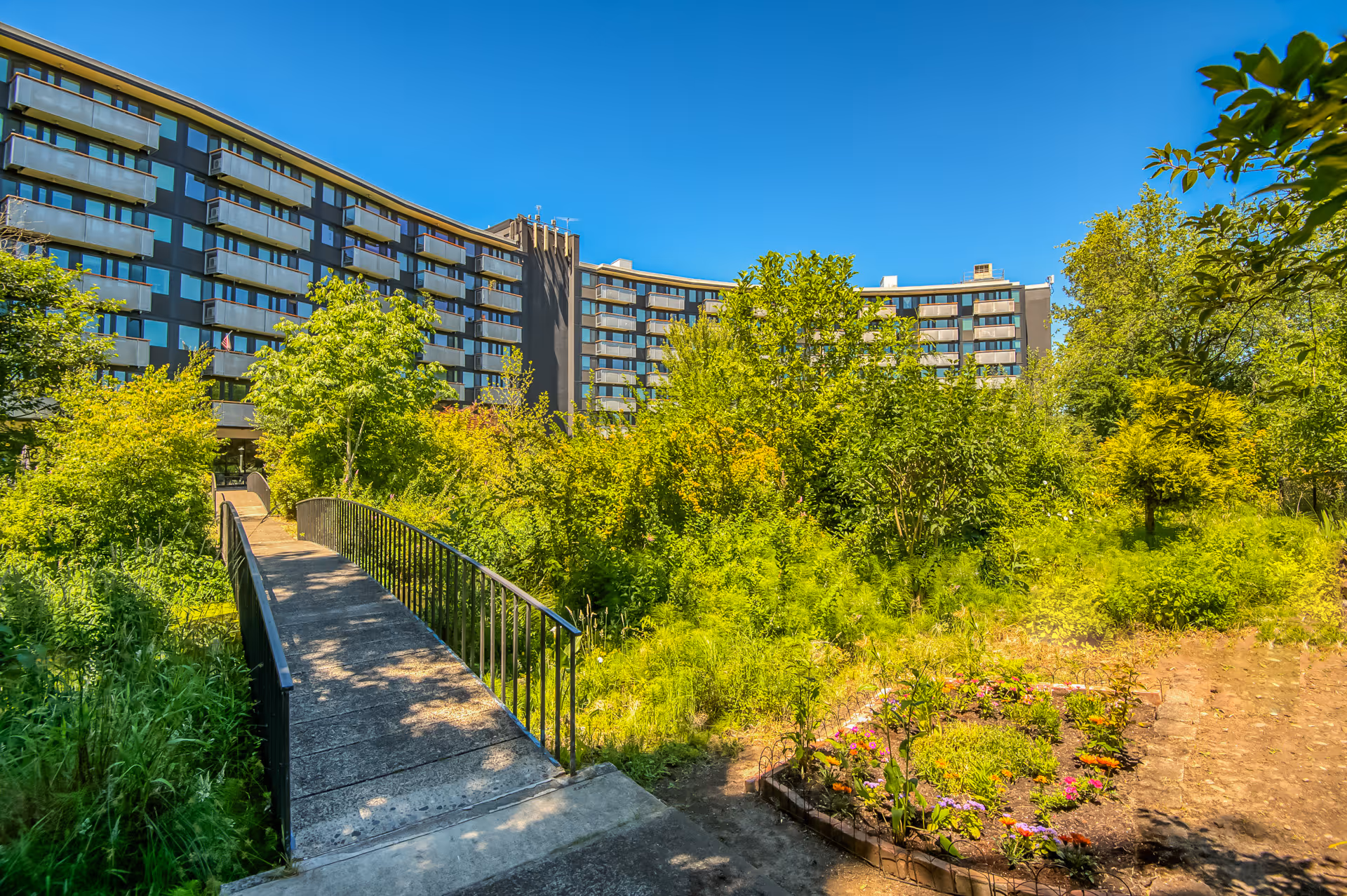 A multi-story senior living facility building with balconies, surrounded by lush green trees and plants under a clear blue sky. A small concrete bridge with metal railings leads into the garden area in front of the building.